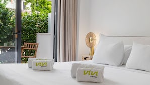 Et hotelværelse med en seng, to hvide håndklæder med 'vivi homes' logo, et natbord med en lampe og udsigt til grønne områder.