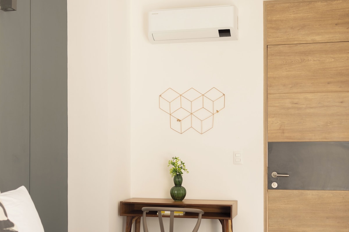 A bedroom with a bed, a desk with a chair, a wall-mounted air conditioner, and a wooden door.