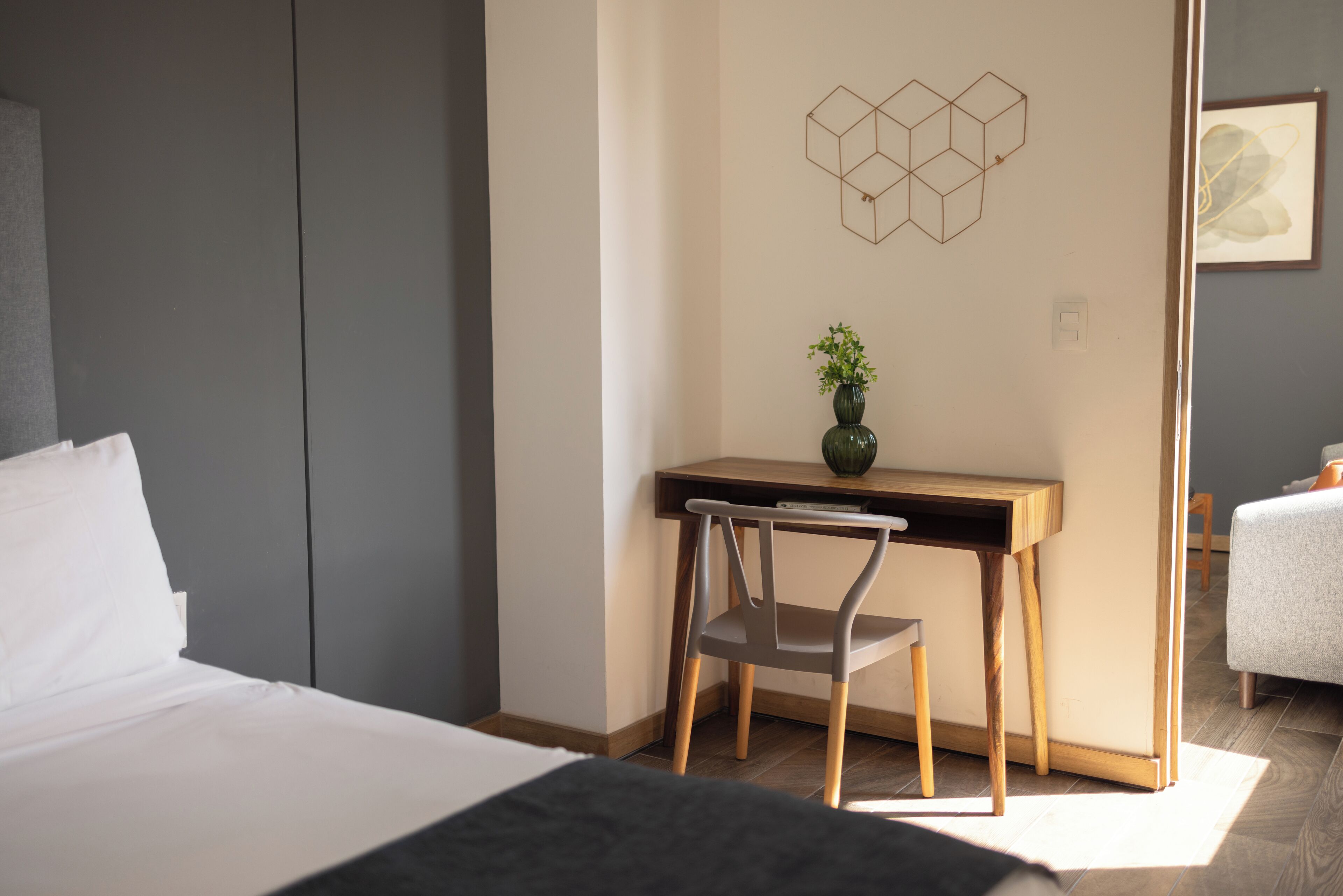 A bedroom with a bed, a desk with a chair, a vase with a plant, and a geometric wall decoration.
