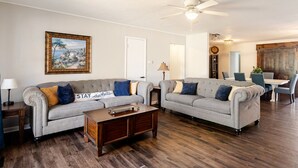 A living room with two grey sofas, a wooden coffee table, a ceiling fan, a dining area with a table and chairs, and a framed painting on the wall. - Magnolia - 4 Bedroom 2 bath & pets allowed (Norman)