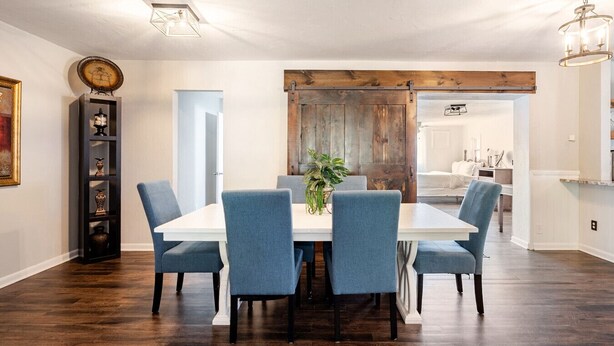 A dining room with a white table and blue chairs, wooden flooring, and a rustic wooden door. - Magnolia - 4 Bedroom 2 bath & pets allowed (Norman)