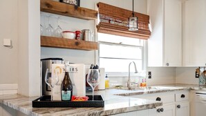 A kitchen with white cabinets, a marble countertop, and a wooden shelf holding wine glasses and a bottle of Asolo Prosecco. - Magnolia - 4 Bedroom 2 bath & pets allowed (Norman)