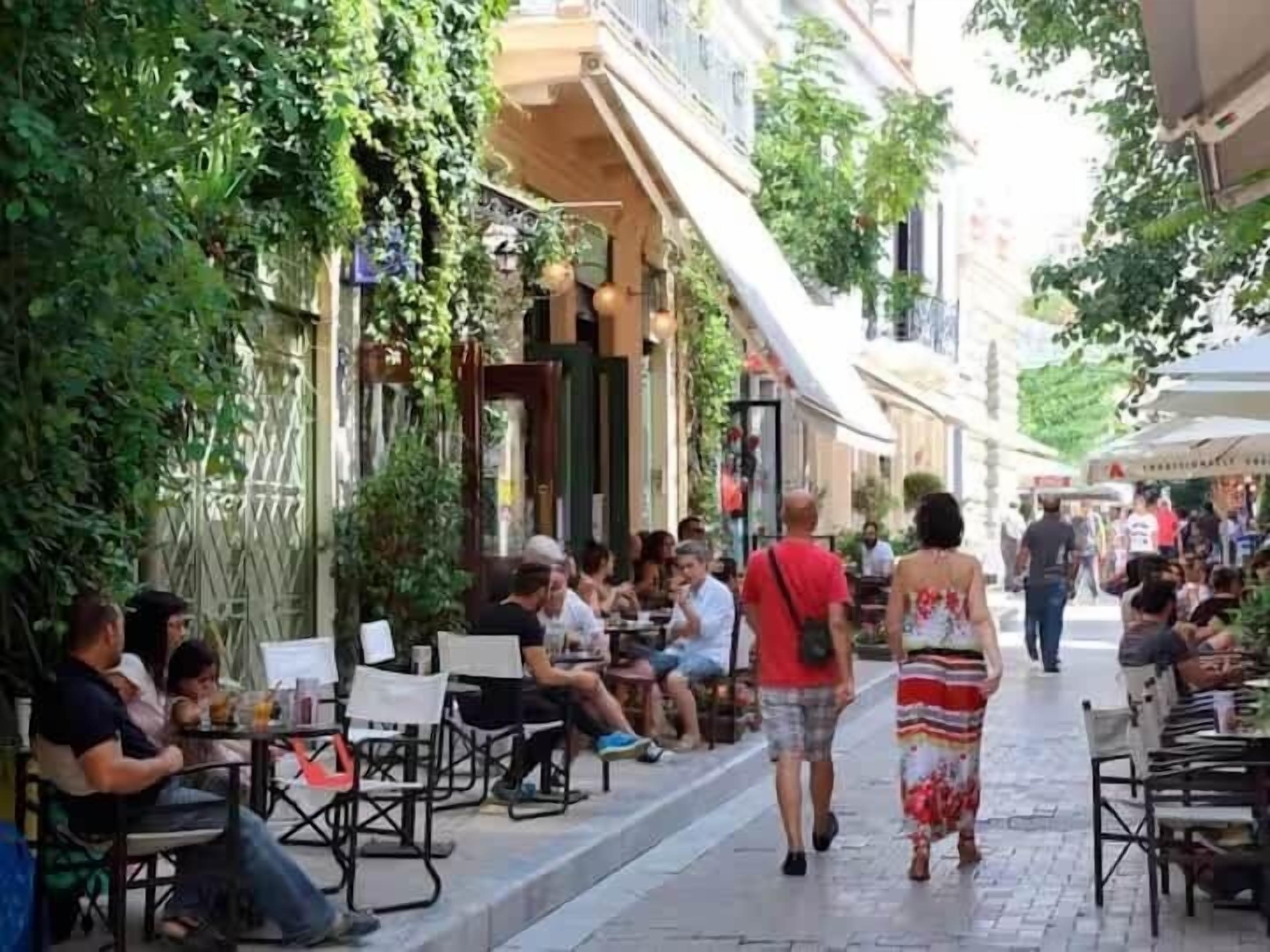 An outdoor café with tables and chairs, surrounded by greenery and buildings.