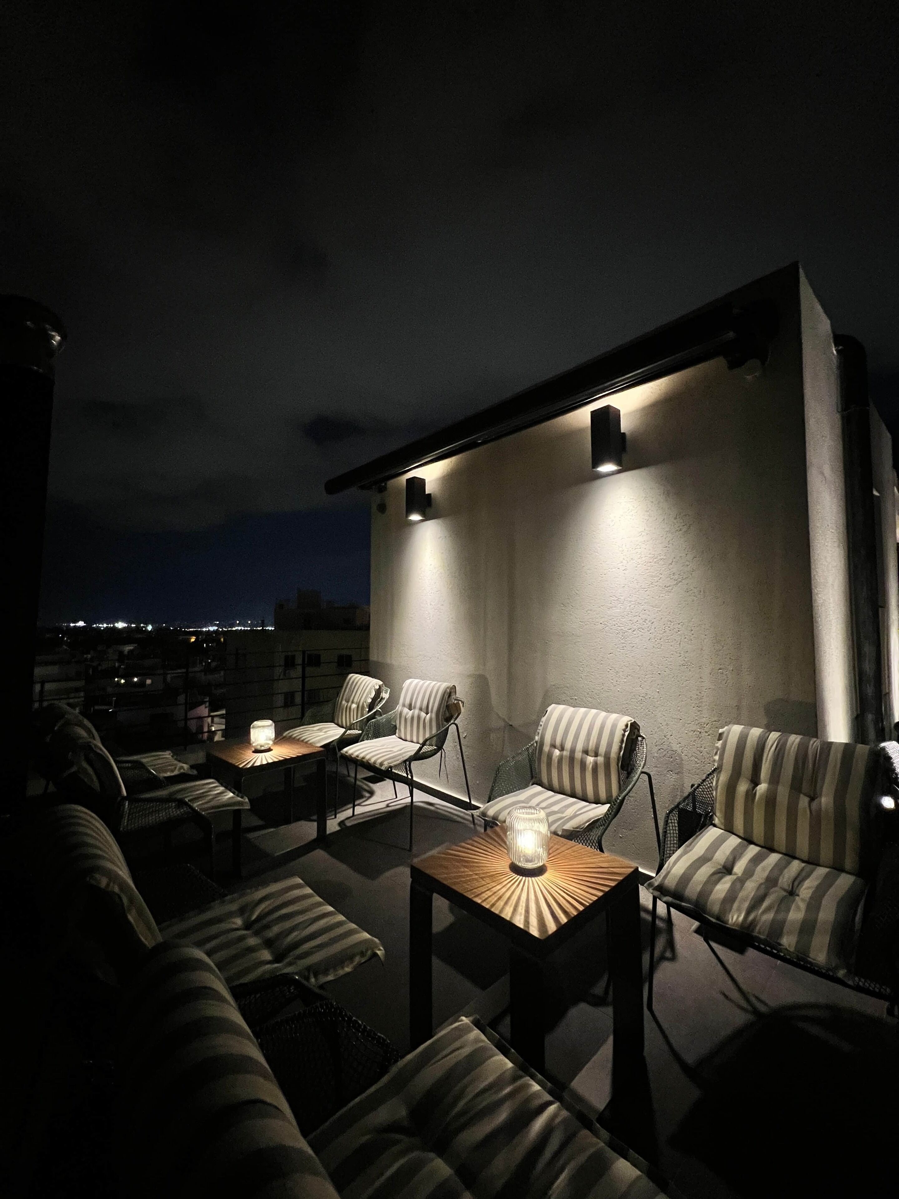 An outdoor seating area with striped upholstered chairs and tables, illuminated by wall-mounted lights.