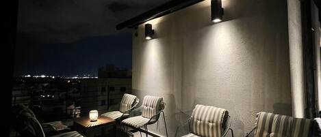 An outdoor seating area with striped upholstered chairs and tables, illuminated by wall-mounted lights.