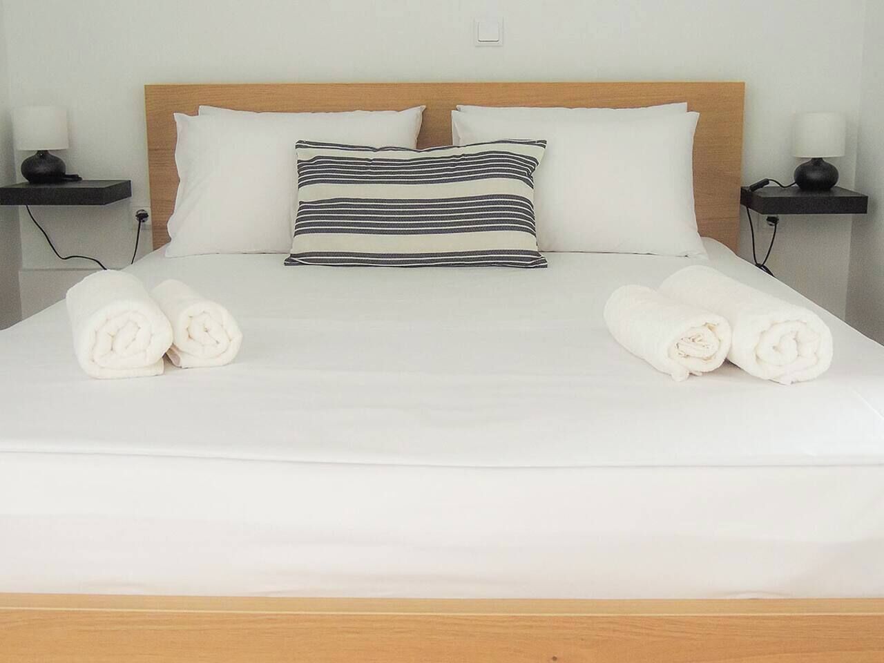 A neatly made bed with white linens, a striped pillow, and rolled towels on the side.