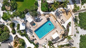 Ein Luftbild zeigt einen Urlaubsort mit einem zentral gelegenen Schwimmbad, umgeben von Palmen und Liegebereichen.