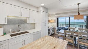 Una cocina con armarios blancos, encimera de madera y vista al océano desde un balcón.