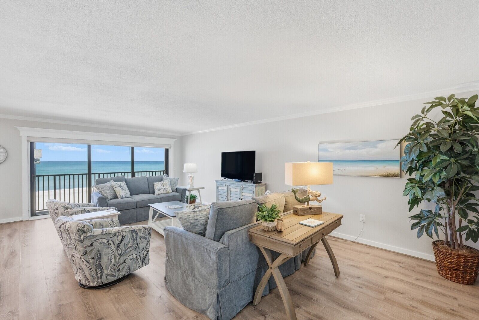 Ein Wohnzimmer mit einer Couch, Sesseln, einem Couchtisch, einem Fernseher und Blick auf den Strand.