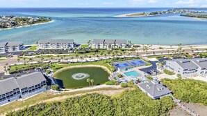 An aerial view of a coastal resort with multiple buildings, a central pool, and a large body of water.