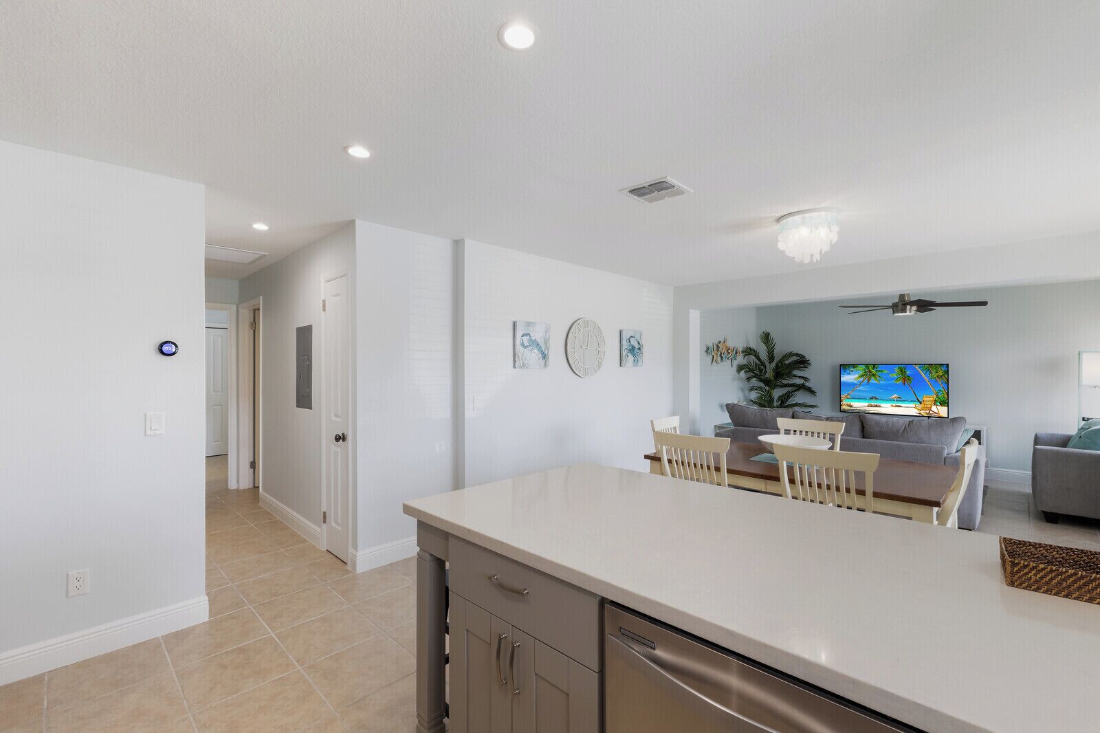 A modern kitchen with a central island, a dining area with chairs, a gray sofa, and a TV displaying a beach scene.