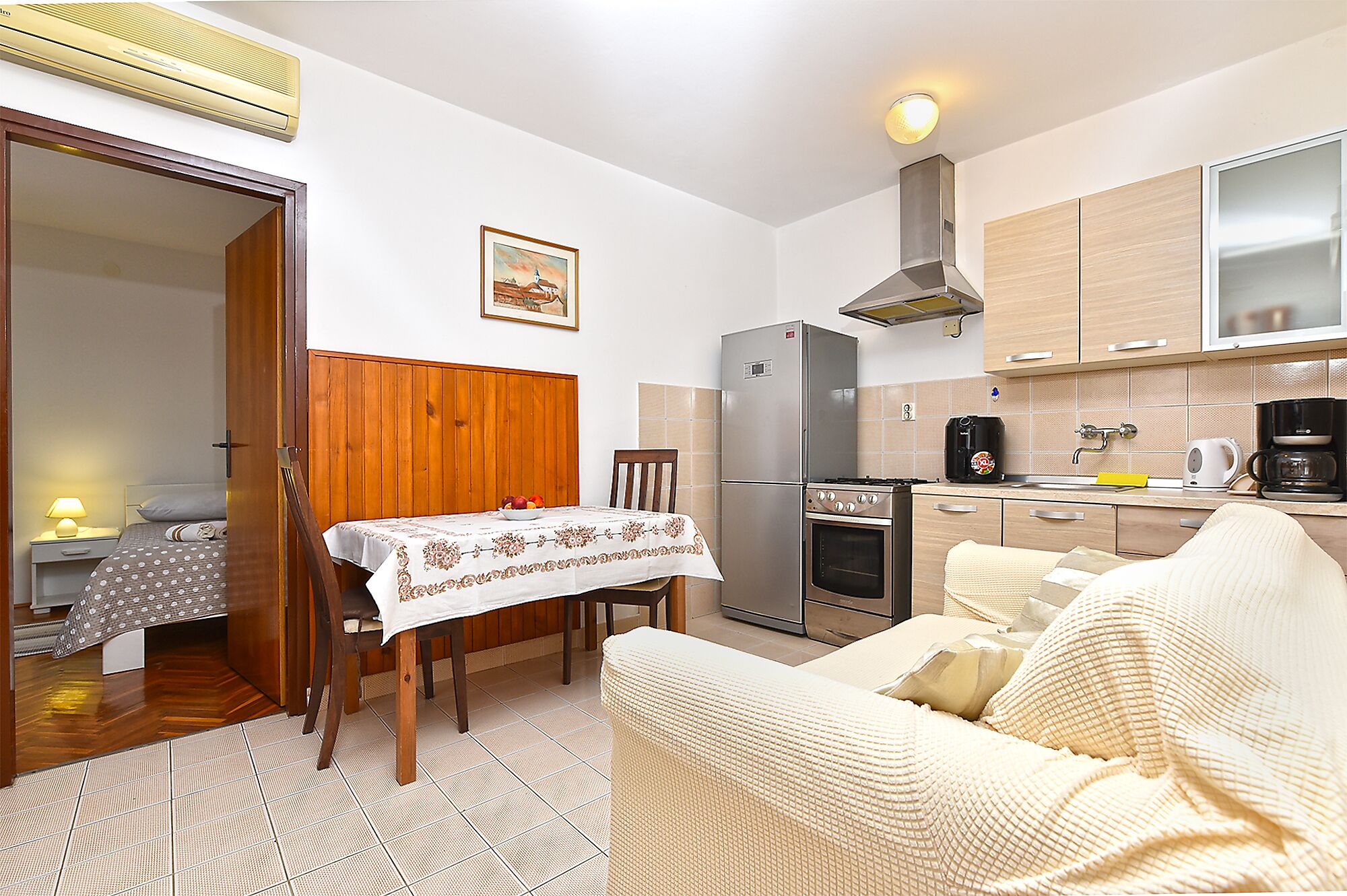 A compact kitchen with a refrigerator, oven, and coffee maker. There's a dining table with a white tablecloth and a chair.