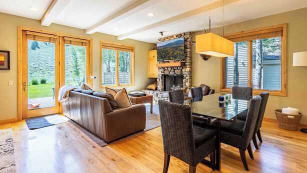 A spacious living area with a large sofa, dining table, and chairs. There is a fireplace with a stone surround and a television mounted above it. Large windows provide natural light and views of the outdoors.