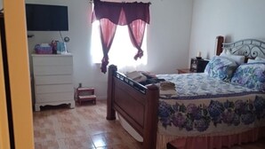 A bedroom with a wooden bed, a white dresser, a television on the wall, and a window with curtains.
