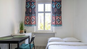 Ein Zimmer mit einem Bett, einem Schreibtisch mit zwei Stühlen, einem Fenster mit Vorhängen und einer Topfpflanze auf dem Schreibtisch.