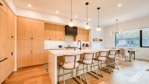 A modern kitchen with wooden cabinets, a central island with bar stools, pendant lighting, and a large window providing natural light.