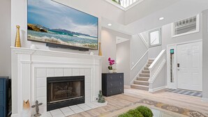 A spacious living room with a fireplace, a large flat-screen TV mounted above it, a staircase leading to another level, and a door with a sign. - Family Villa | Deck, Lagoon Views, Complex Pool, Bikes (Hilton Head Island)