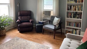 Een woonkamer met een leren fauteuil, een grijze fauteuil, een boekenkast en een potplant.
