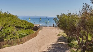 Un sentier sablonneux qui mène à une plage où l’on peut voir des voiliers et des pratiquants de planche à voile.