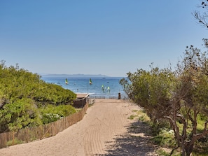 A sandy pathway leading to a beach with sailboats and windsurfers.