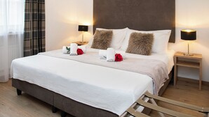 A neatly made bed with white linens, two fluffy pillows, and a wooden headboard. There are two bedside tables with lamps, and a wooden chair is visible in the foreground.