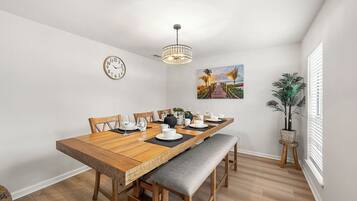 Un coin repas avec une table en bois dressée pour six personnes, un banc, un lustre, une horloge et un tableau accroché au mur.