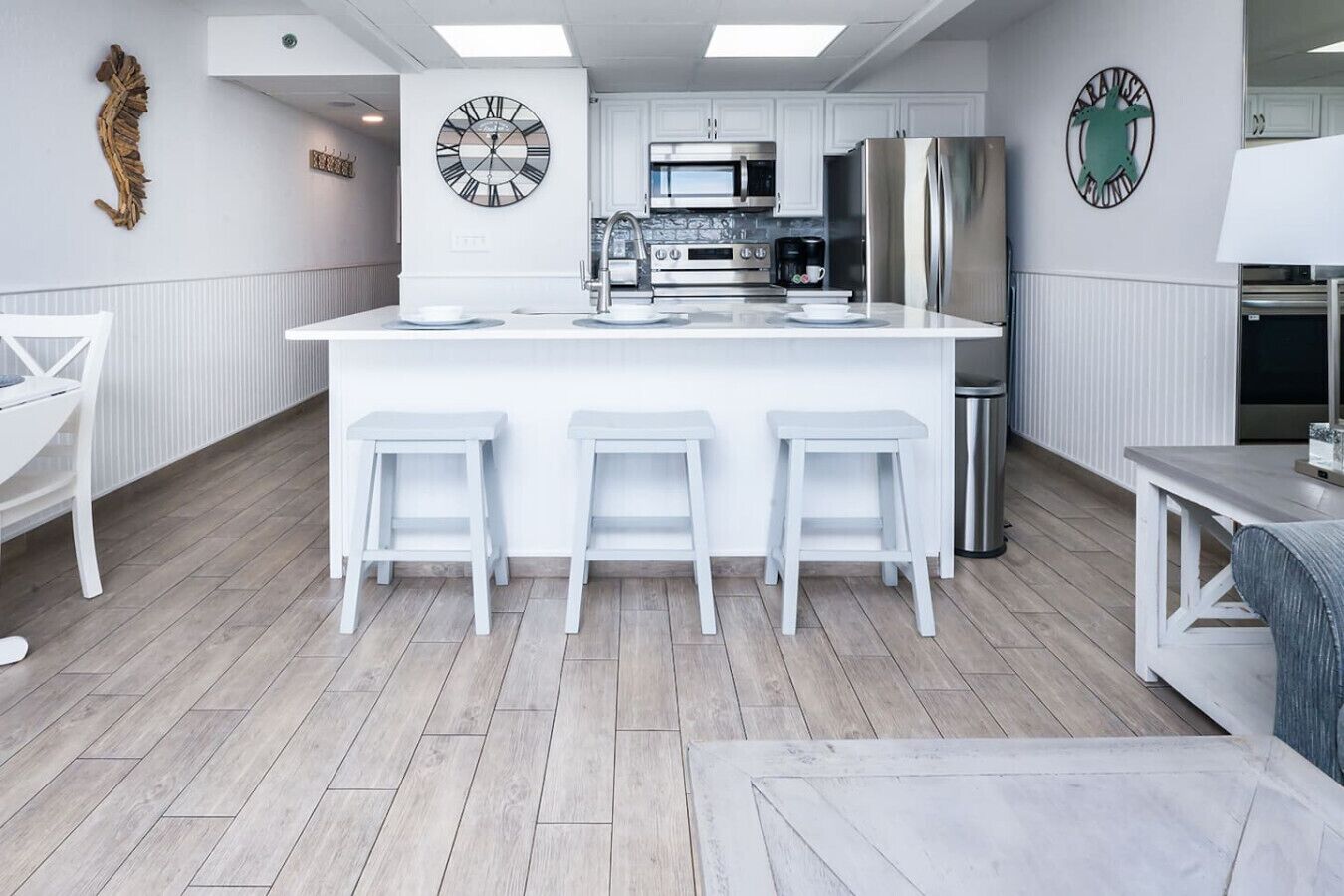 Una cocina moderna con armarios blancos, una isla central con taburetes, electrodomésticos de acero inoxidable y relojes decorativos en la pared.