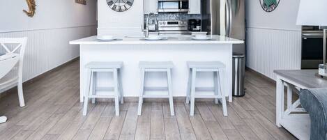 Una cocina moderna con armarios blancos, una isla central con taburetes, electrodomésticos de acero inoxidable y relojes decorativos en la pared.