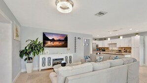 A modern living room with a large sectional sofa, a dining area with a kitchen in the background, and a large landscape painting on the wall. - Barefoot In Lantana Paradise (Lantana)