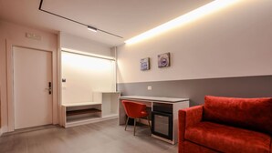 Ein modernes Hotelzimmer mit Schreibtisch, einem roten Sessel, einer roten Couch und einem kleinen Schrank.
