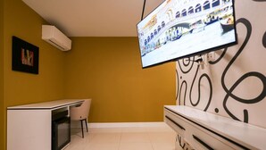 Ein Hotelzimmer mit an der Wand montiertem Fernseher, der eine Stadtansicht zeigt, einem Schreibtisch mit Stuhl und einer Minibar.