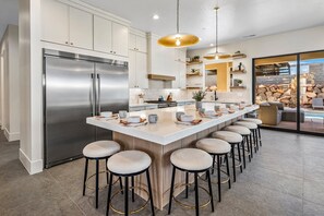 Una cocina moderna con refrigerador de acero inoxidable, una isla central con taburetes de bar, iluminación colgante y vista a una pared de piedra.