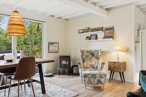 Uma sala de estar moderna com piso de madeira, uma área de refeições com cadeiras, uma poltrona estampada, uma prateleira fixada na parede com quadros e uma lareira a lenha.