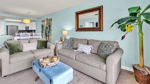 A living room with two sofas, a coffee table, a ceiling fan, a kitchen in the background, and a large mirror on the wall.