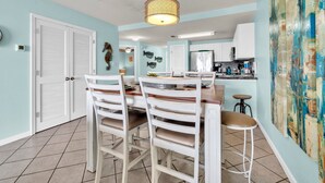 A dining area with a wooden table and white chairs, a hanging light fixture, and a kitchen in the background.