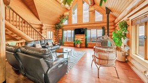 A spacious living room with a black leather sofa, a round coffee table, and a wicker chair. There is a large window with a view of the outdoors.