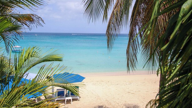 A beach with clear turquoise water, a sandy shore, and a few lounge chairs under a blue umbrella.