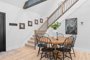 Un comedor con mesa de madera y sillas de metal negro, una escalera con barandillas de madera y cuadros en la pared.