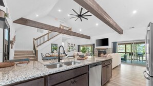 A spacious kitchen with granite countertops, a dishwasher, and a large island. There is a staircase leading to an upper level, and a large window providing a view of the outdoors.
