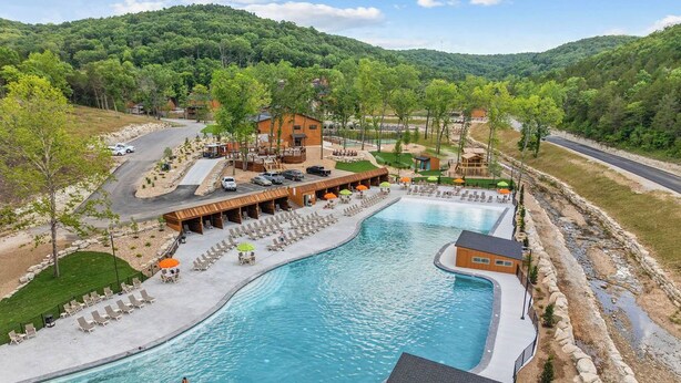 A large outdoor swimming pool with lounge chairs and umbrellas, surrounded by a building with a deck and a view of a forested hill.