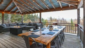 A covered outdoor seating area with a long wooden table, cushioned seating, and string lights. - Moose Ridge Haven | Spacious and beautifully appointed home | Hot tub & Sauna (Rangeley)