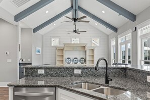 Una cocina con encimeras de granito, un lavamanos de acero inoxidable, ventiladores de techo y vistas al área de estar a través de amplias ventanas.