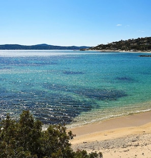 Une plage à l’eau turquoise et limpide, un rivage sablonneux et, au loin, une colline parsemée de bâtiments.