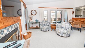 Un salon avec une cheminée, deux fauteuils, un canapé, une table à manger avec des chaises, et une vue sur l’extérieur grâce à de grandes fenêtres.