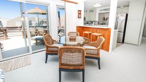 Un salon moderne avec une table en verre entourée de chaises en osier, un bar en bois et un coin cuisine doté de meubles blancs et d’un réfrigérateur.