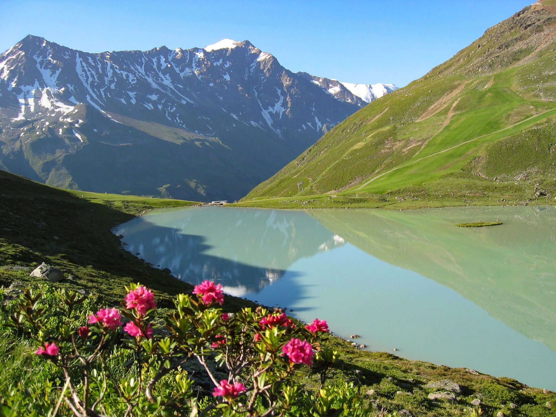 A mountain lake with a clear reflection, surrounded by green hills and a snow-capped mountain in the background.