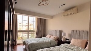 A bedroom with two beds, a wardrobe, a window with curtains, an air conditioning unit, and a bedside table with a lamp.