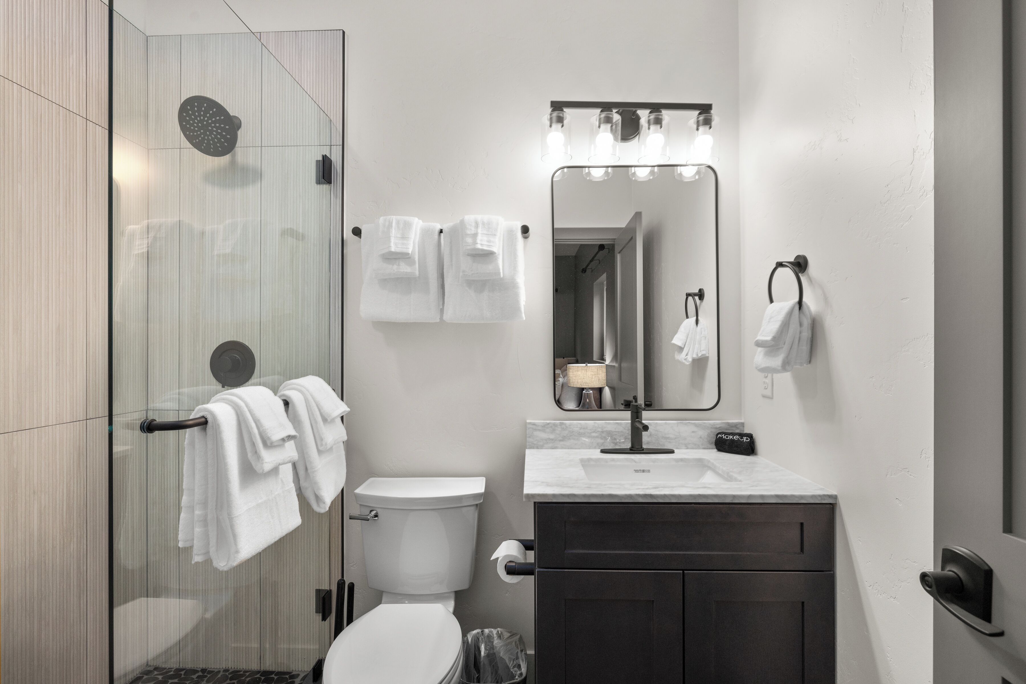 A modern bathroom with a glass shower enclosure, a white toilet, a vanity with a mirror, and towels on hooks.