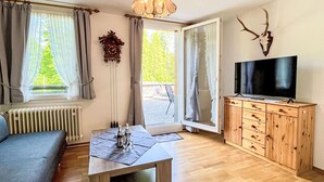 Un salon comprenant un canapé, une table en bois avec des verres, un téléviseur posé sur un meuble en bois, et une grande fenêtre avec des rideaux.
