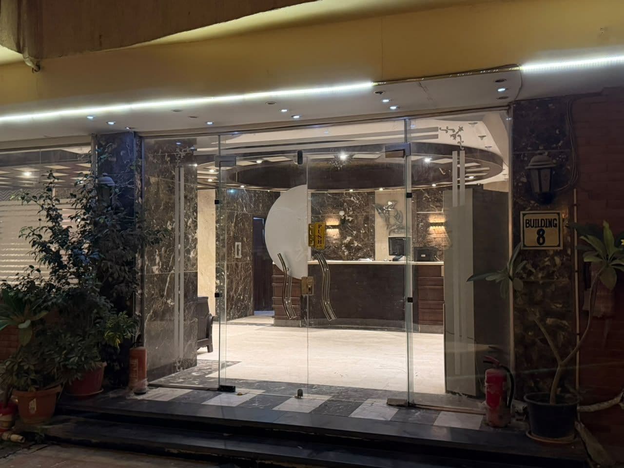 Entrada de hotel con puerta de vidrio, mostrador de recepción y un cartel con el número del edificio.